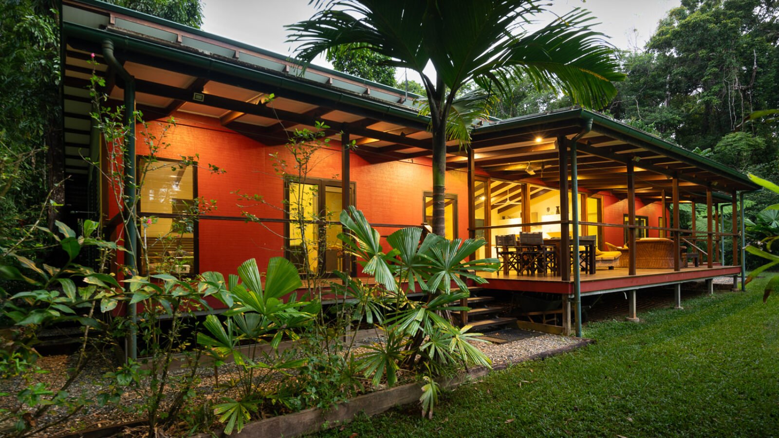 The House at dusk, verandah lit, rainforest behind