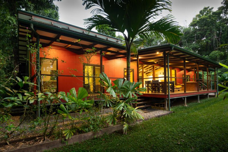 The House at dusk, verandah lit, rainforest behind